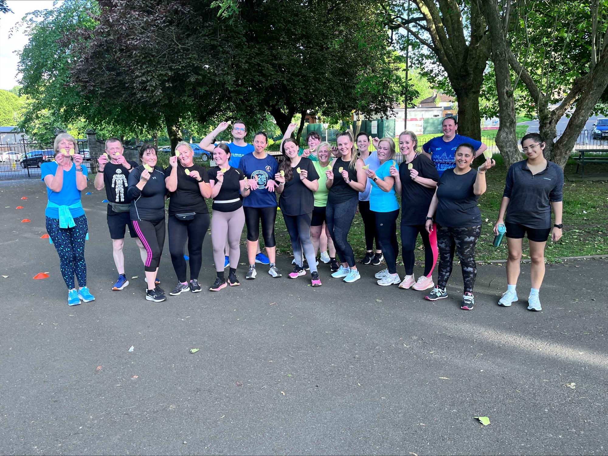 A group of twenty individuals, mostly women, smiling and posing in a park, wearing workout clothes and holding weights. They stand in front of trees, with a clear blue sky in the background.